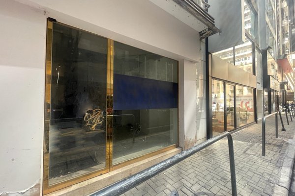 Ref#3251: Vacant storefront with large windows, a gold-framed glass door, and a blue horizontal window stripe, located along a city sidewalk with nearby buildings and metal railingideal for shop or office rental.