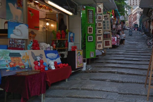 Ref#3583 features a narrow stone staircase bordered by outdoor art stalls with vibrant paintings and red figurines, bustling with visitorsperfect for anyone seeking to rent a shop in an artistic environment.