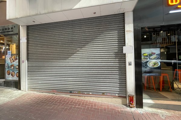 The entrance of Ref#3183, a city shop for rent, is secured by a closed metal roller shutter. Next door on the right, a restaurant displays food photos and orange chairs along the brick sidewalk.