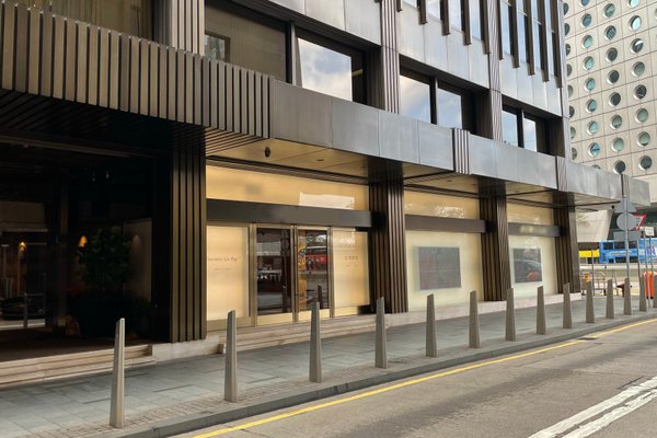 Street view of Ref#3130, a modern building with tall windows and metallic vertical beams—ideal for office rental. A two-lane road and short bollards line the sidewalk in front. No people are visible.