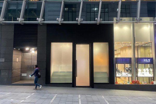 A person walks by the modern facade of Ref#3199, featuring a black entrance and large frosted glass door—prime rental shop space—situated next to a brightly lit jewelry store window showcasing jewelry.