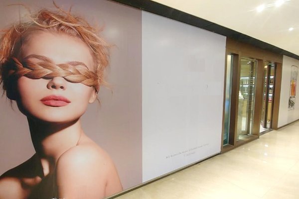 A large wall poster in a shopping mall advertises Ref#3418 and shows a calm-faced woman with nude makeup and light skin, her short tousled blonde hair braided around her eyes like a blindfold.