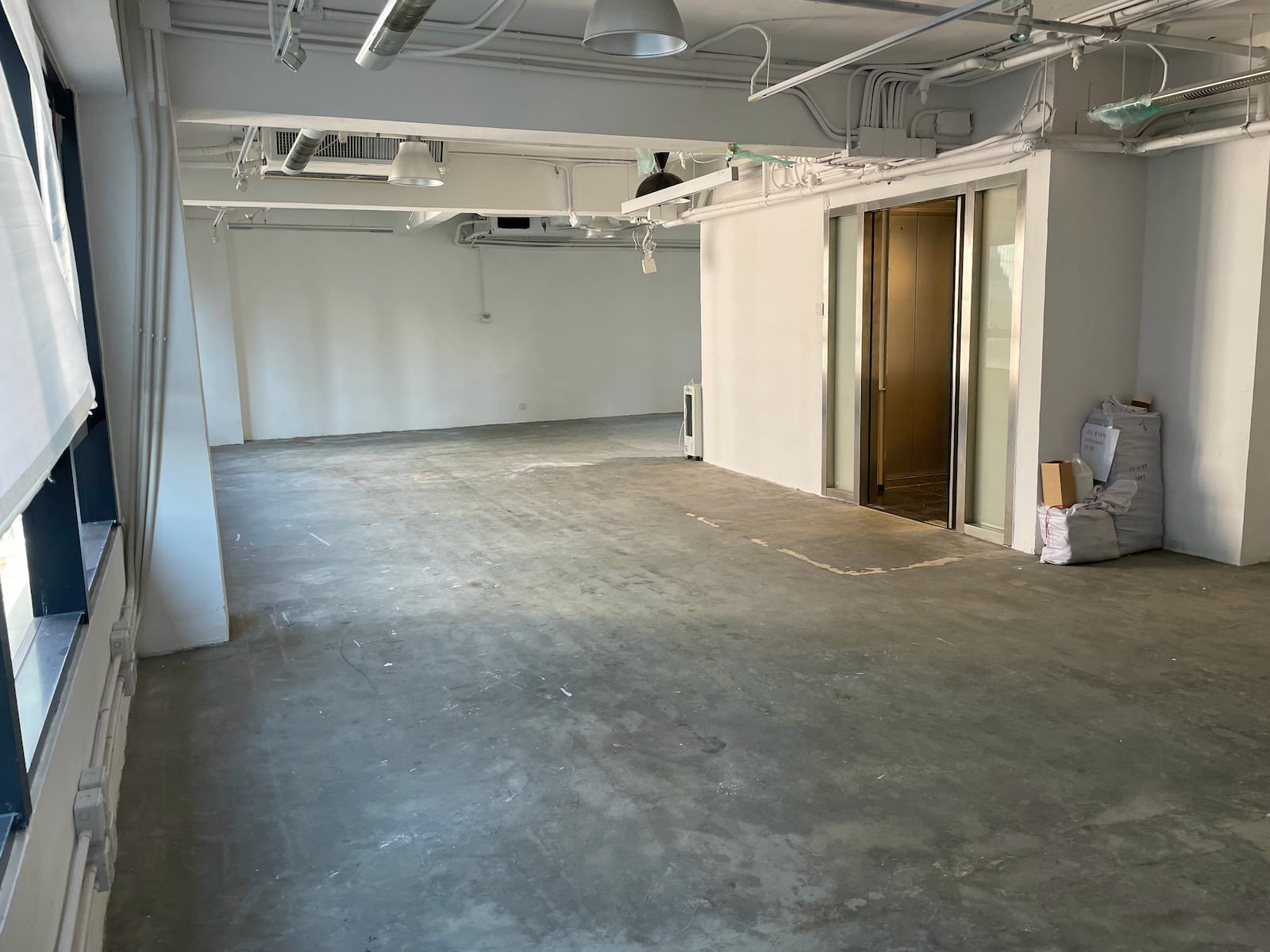 Ref#3582: Industrial-style space with concrete floors, white walls, exposed ceiling pipes, and large windows on the left. Elevator doors and stacked boxes are on the right; ideal for restaurant or office rental.