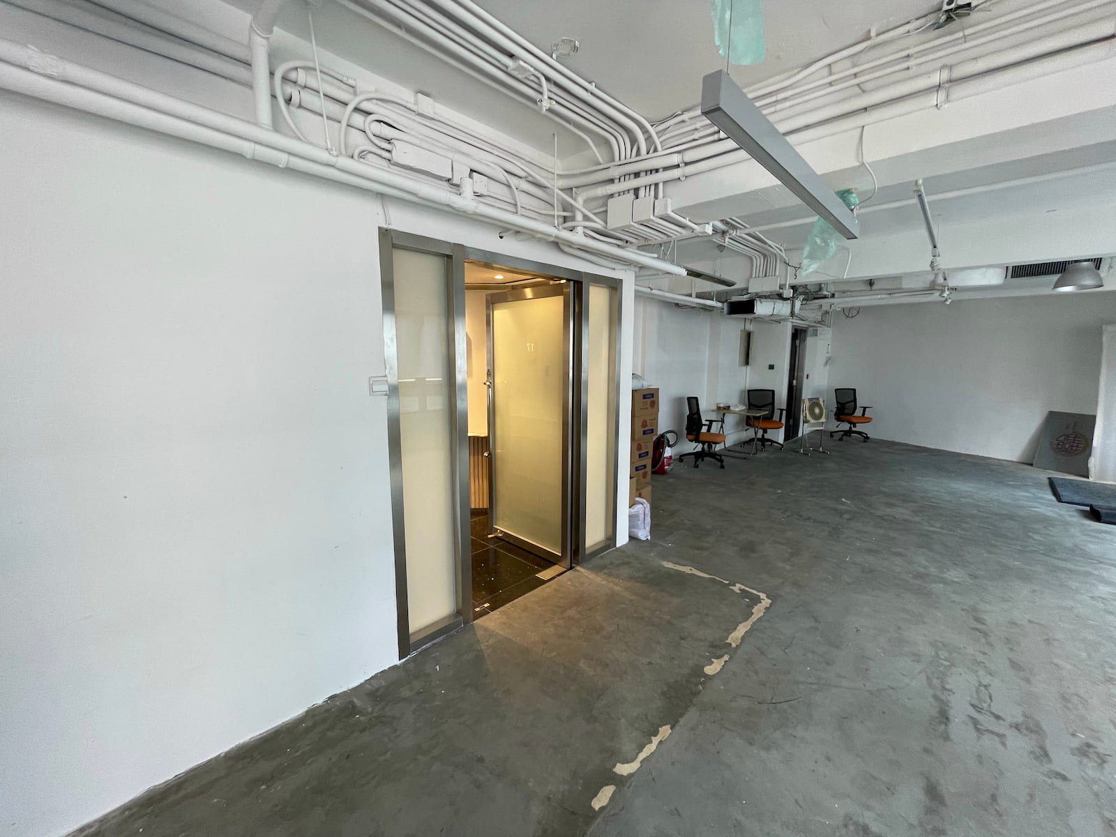 Ref#3582: A mostly empty office with an open elevator door, scattered chairs, exposed ceiling pipes, and unfinished flooring12ideal for anyone seeking to rent office space with a modern industrial aesthetic.