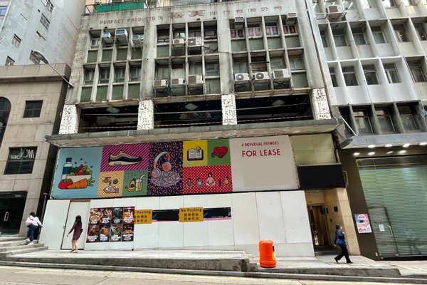 Street view of vacant storefront (Ref#3239) in city, featuring colorful artwork panels, posters, and a large For Lease sign 6great spot to rent for a shop or restaurant. A few pedestrians pass by on the sidewalk.