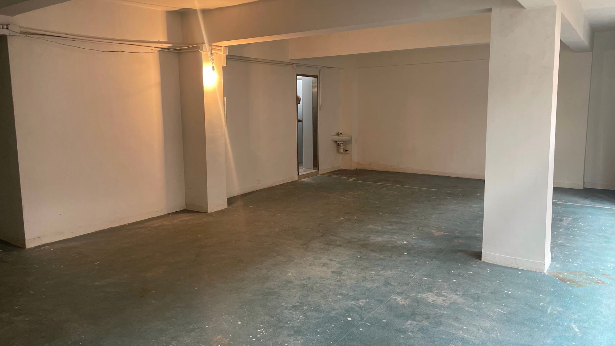 Ref#3587: A mostly empty room with white walls, exposed wiring, support columns, a water fountain at the back, and a worn floor1ideal for rent as a shop or office. Features include a closed rear door and left-wall light fixture.