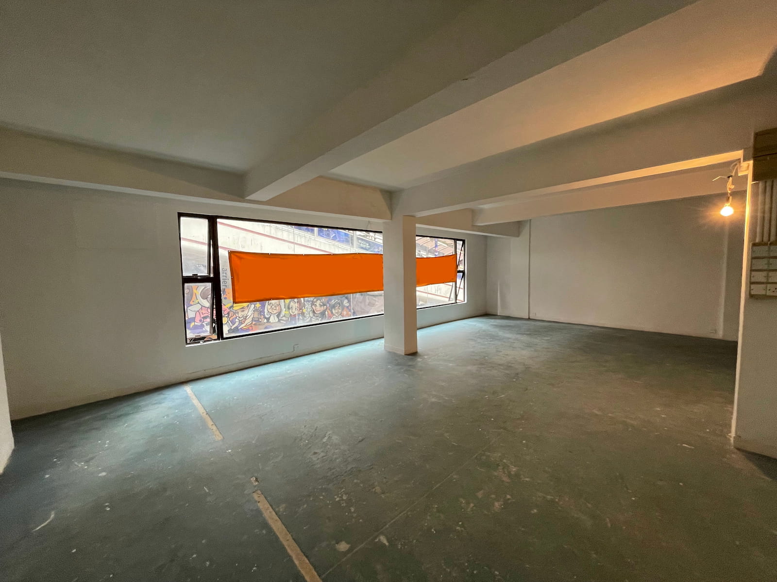 Ref#3587: Industrial-style space with concrete floors, white walls, exposed beams, and large windows with orange panels. Ideal for shop or office rental. Natural and artificial light; outdoor graffiti visible from inside.