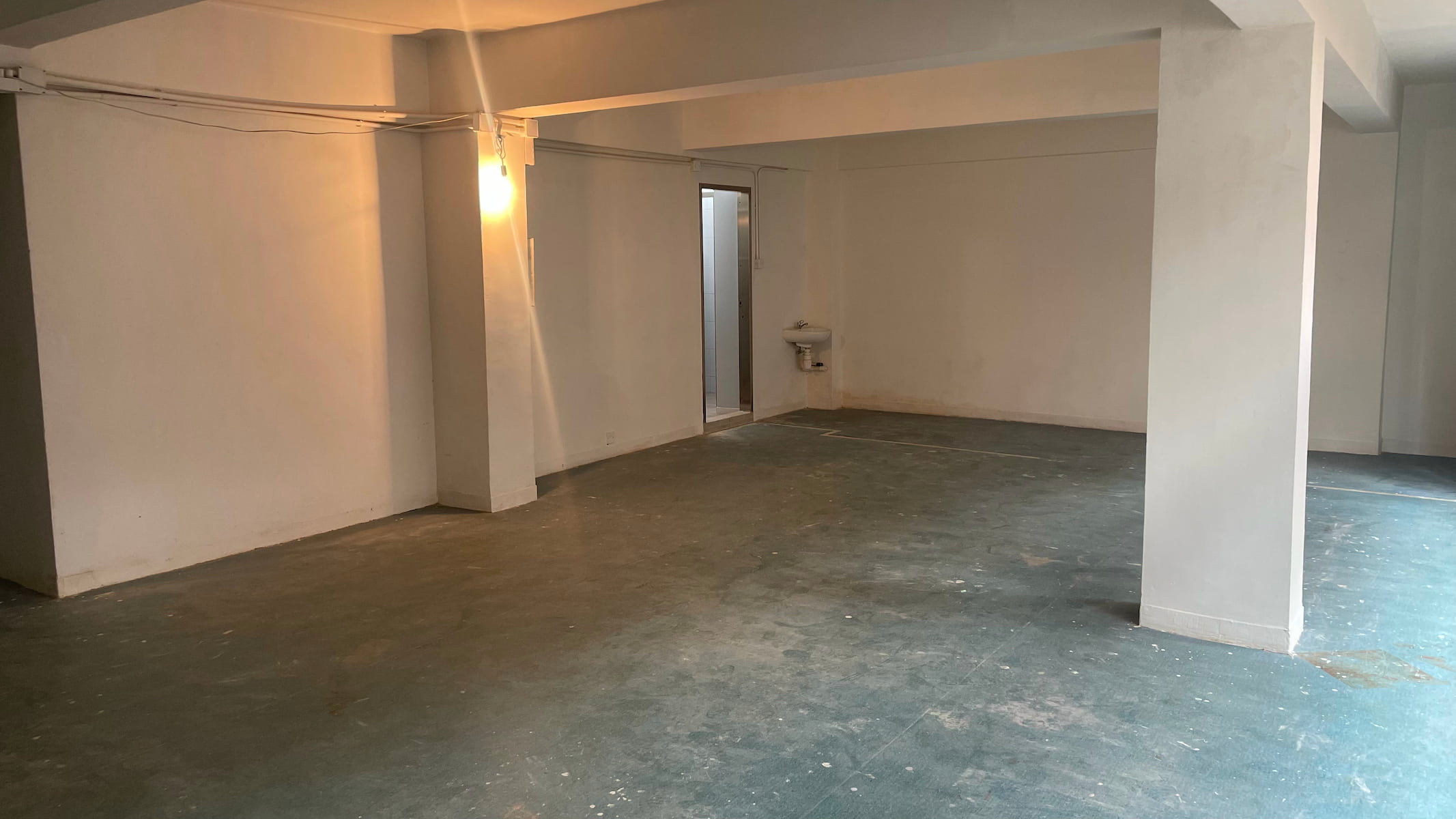 Ref#3587: Mostly empty, unfinished space with white walls, exposed wiring, single light, water fountain in the corner, and concrete floor1ideal for renting as an office or shop and customizing to suit your needs.