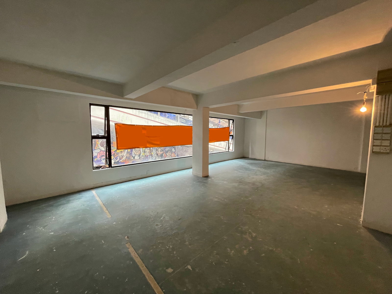 Ref#3587: An empty, unfurnished space with white walls, exposed beams, and a green floor1ideal for rent as an office or shop. Features large windows with orange coverings and a single hanging light bulb.
