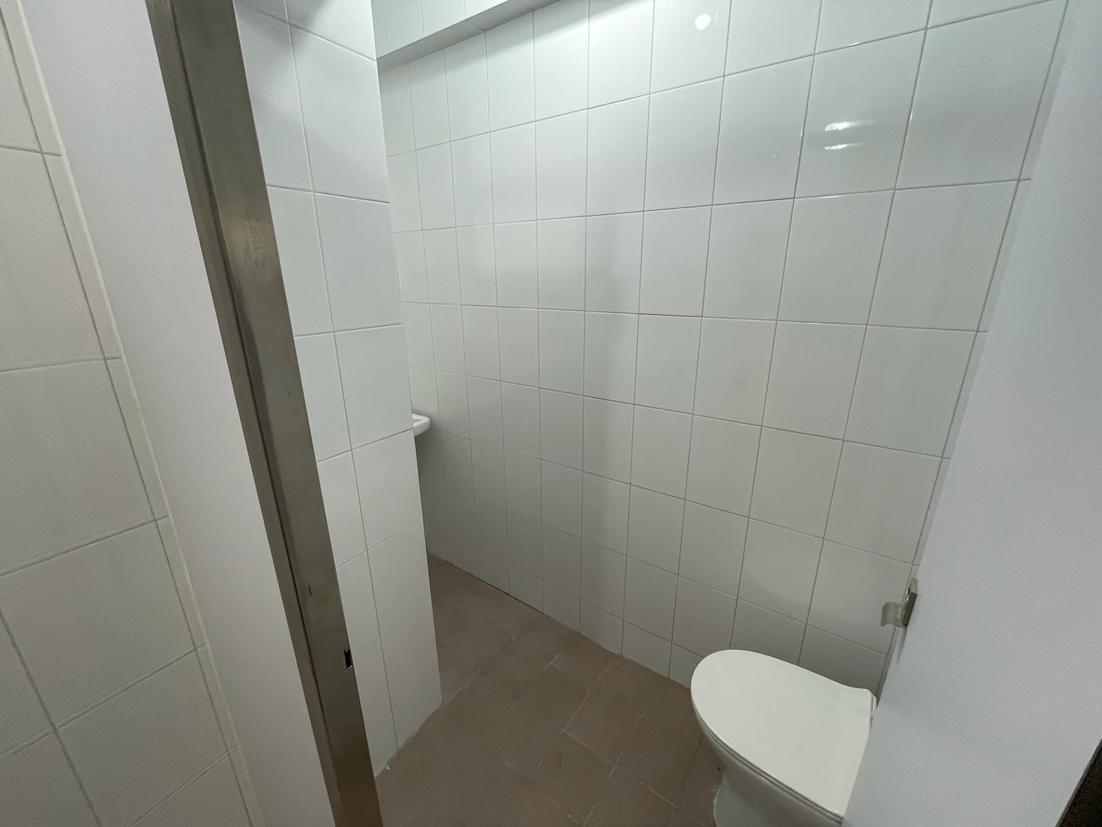 Ref#3587: Compact bathroom with white tiled walls, light brown floor tiles, white toilet, and a partial view of a corner sink1perfect minimal facilities for rent shops or offices, separated by a partial wall.