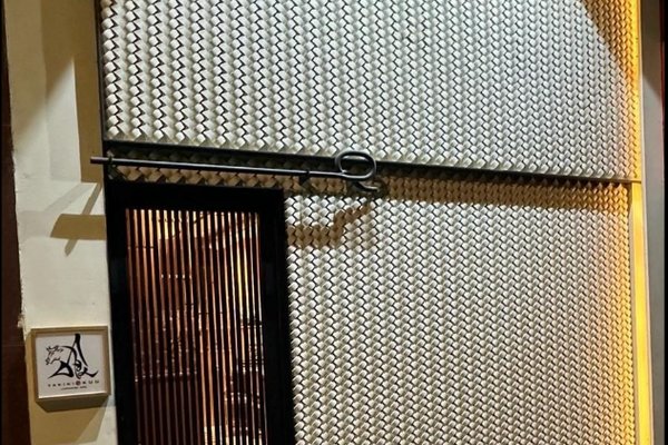 Ref#3461: Building facade with a sliding metal gate in geometric pattern beside a vertical-barred door. Warm interior lighting makes it ideal for restaurant rental. A sign with stylized black writing and drawing hangs on the wall.