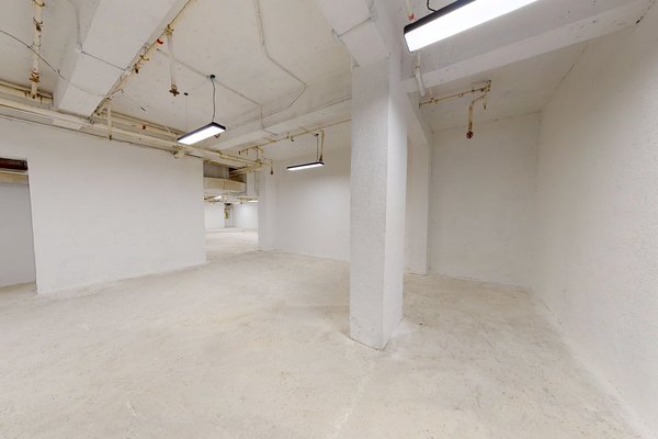 Ref#3136: Spacious empty basement with white walls, exposed ceiling pipes, fluorescent lighting, and central pillar—perfect for rent as a shop or office.