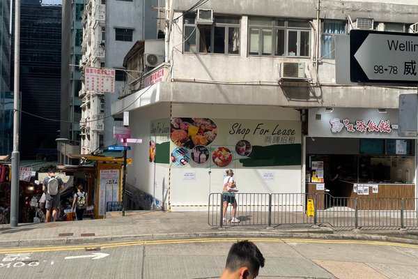 A woman stands before Ref#3132, a shop with a "For Lease" sign on a city corner. Next to it, a restaurant with Chinese signage attracts passersby who stroll the sunny sidewalk.