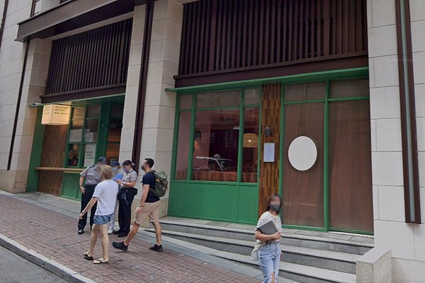 A group converses on a sidewalk by a building with large green-framed windows and doorsRef#3485, ideal for office or shop rentalwhile one person stands alone in front.