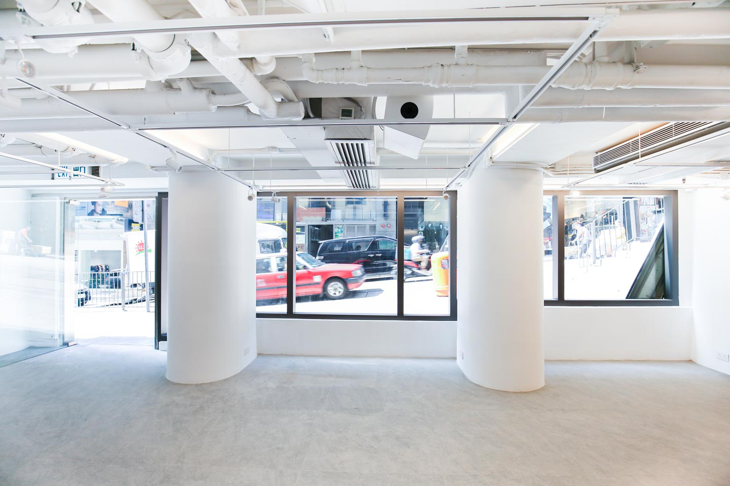 Ref#3489 is a bright, modern office rental with large windows, exposed white ceiling pipes, and two round columns. The empty interior is filled with natural light and offers views of cars and pedestrians outside on a sunny day.