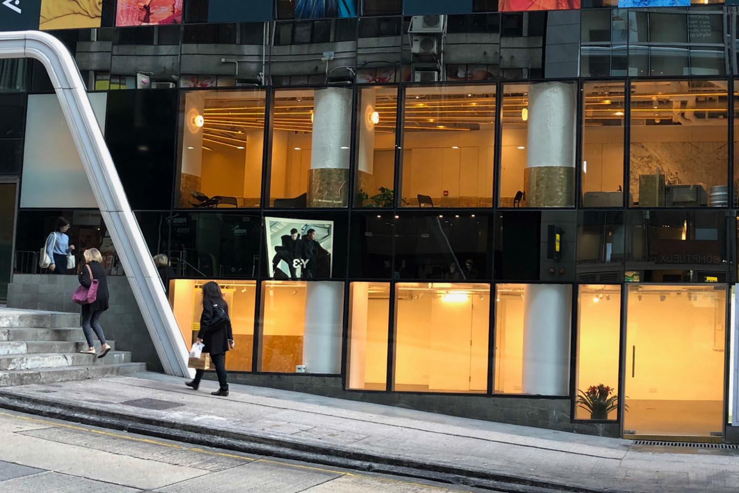 Street view of a modern glass building with two floors lit up; people walk past the steps. A digital screen displays three people and the EY logo, promoting Ref#3489 prime shop and office rental opportunities.