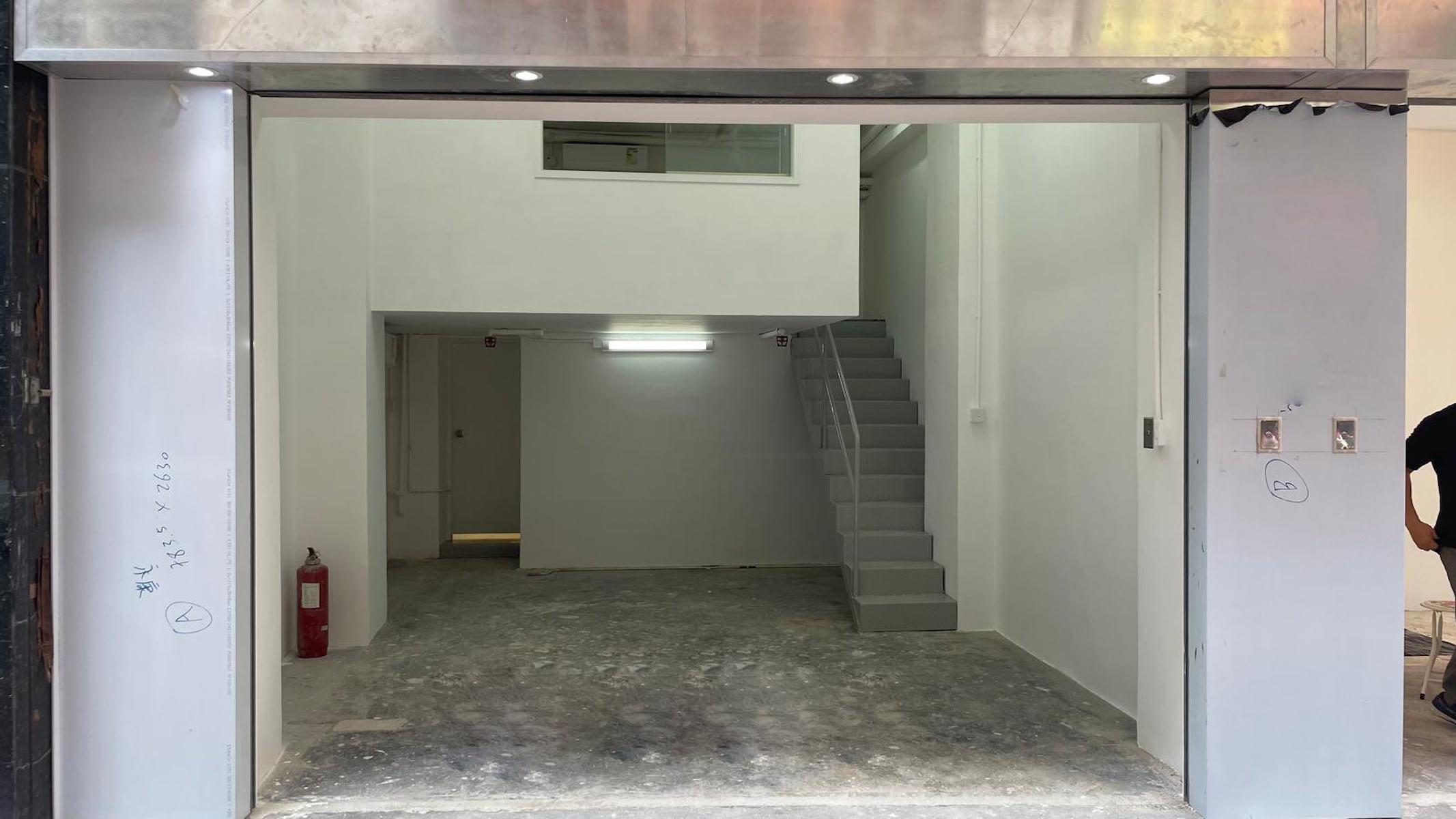 Ref#3038: Unfinished commercial space for rent, ideal as office or shop, with white walls, exposed concrete floor, gray staircase on the right, fire extinguisher to the left, and a small upper-level area.
