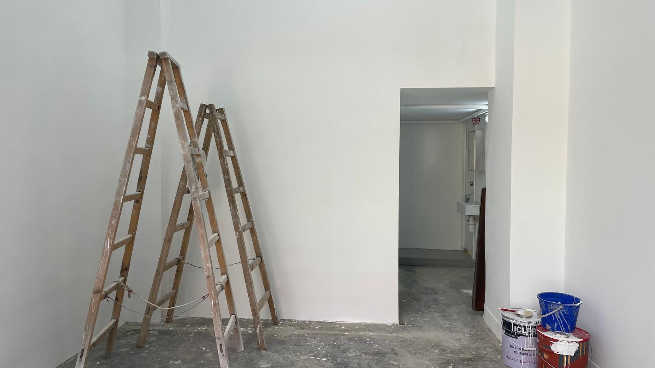 Ref#3039: Two wooden ladders stand against a freshly painted white wall in an empty room with a concrete floor
perfect for a shop or office. Buckets and paint supplies are in the corner, and an open doorway leads to another room.