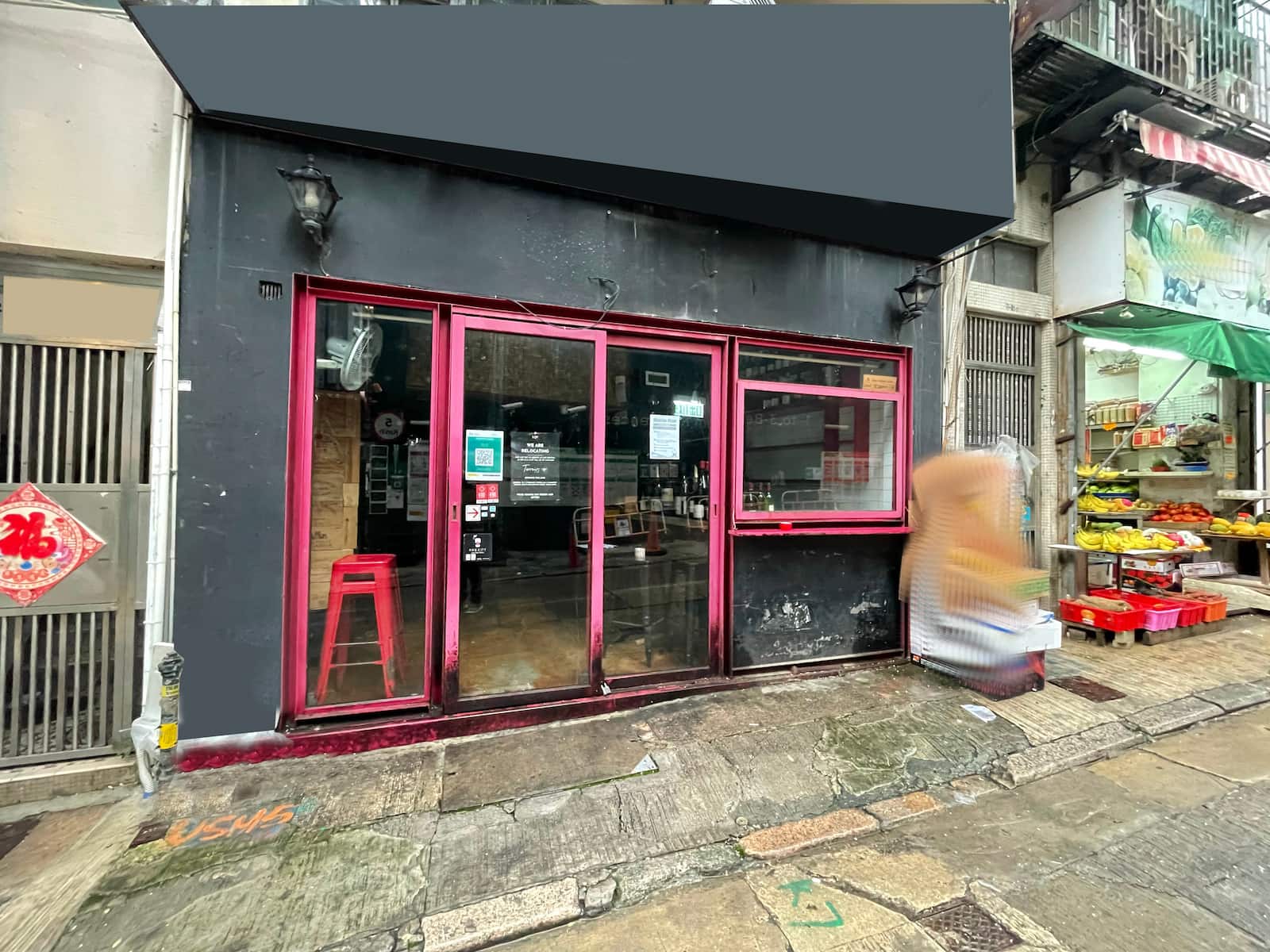 Ref#3036 is a small storefront with black walls, red trim, glass doors and windows, ideal for shop rental. Inside is a red stool; outside, fruits and vegetables are displayed on the right. A pedestrian walks by on the sidewalk.