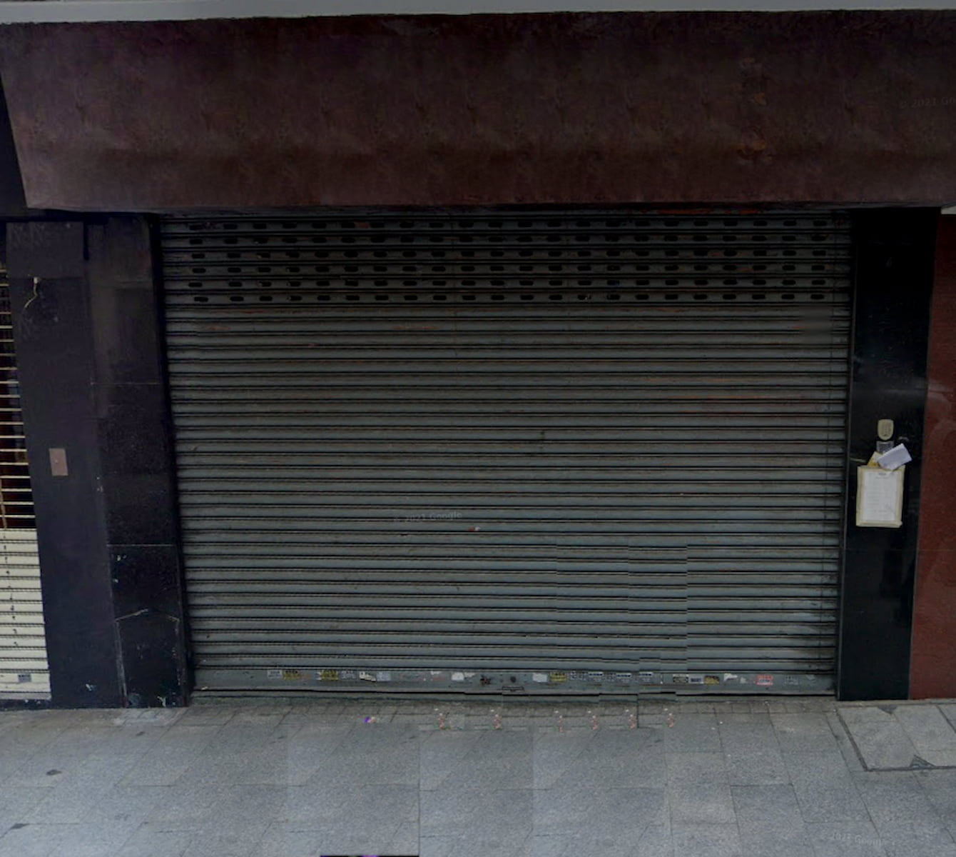 Ref#3037: Metal security shutter is fully closed over a storefront, hinting at a rental opportunity. A clipboard or paper is posted on the right side of the entrance, and the empty sidewalk lies in front.