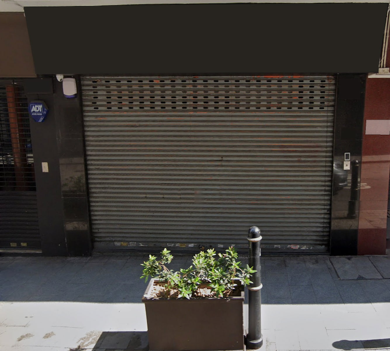 A metal roller shutter covers the storefront of Ref#3037, which is available to rent. Above, a blank black sign is ready for your branding. Outside, a rectangular planter with green plants and a short black bollard are on the paved sidewalk.