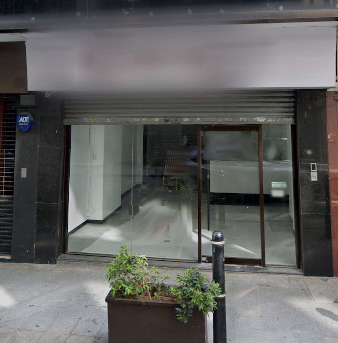 Ref#3037: Storefront with large glass windows and a partially open metal shutter, ideal to rent for a shop or office. The empty interior awaits your vision, with a planter of green plants by the entrance.
