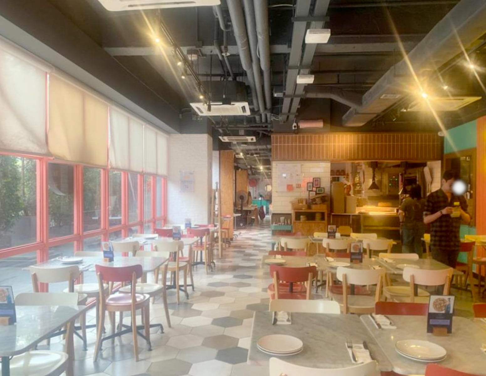 Ref#3029 features a bright, modern restaurant with hexagonal tile floors, red-framed windows, and tables set for dining
making it ideal for restaurant rentals. A few people are working near the kitchen counter in the background.