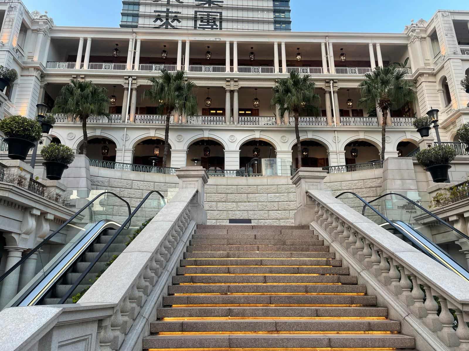 Ref#3013 features a grand staircase with yellow-edged steps leading to a white, multi-story building with arched balconies, columns, palm trees, and decorative lampsideal for renting office or shop space in an elegant setting.