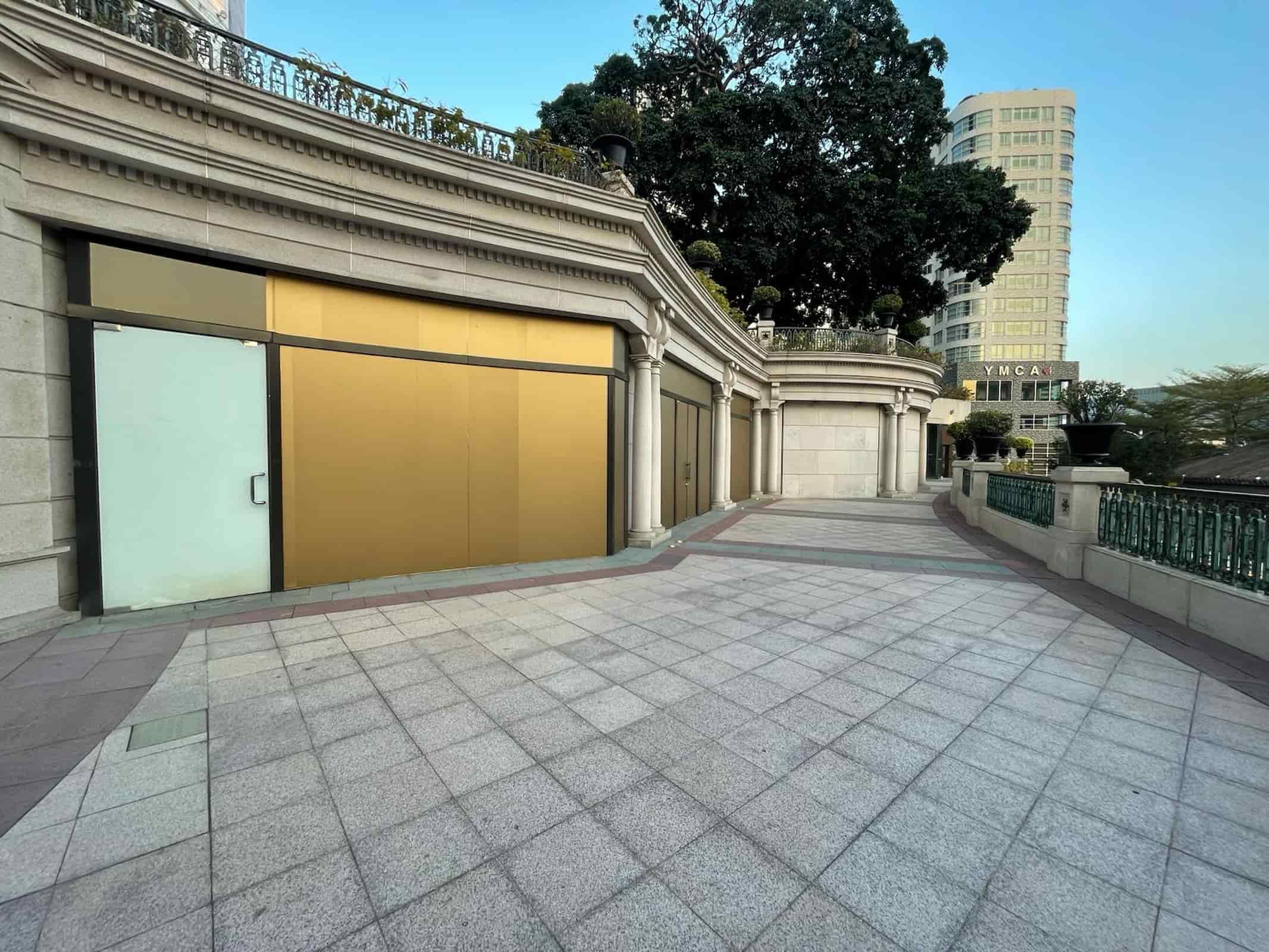 An empty, tiled walkway curves past closed storefronts with gold and white panelsideal rental spots for shops or restaurants. In the background, trees and a tall building marked "YMCA" are visible. Product: Ref#3013.
