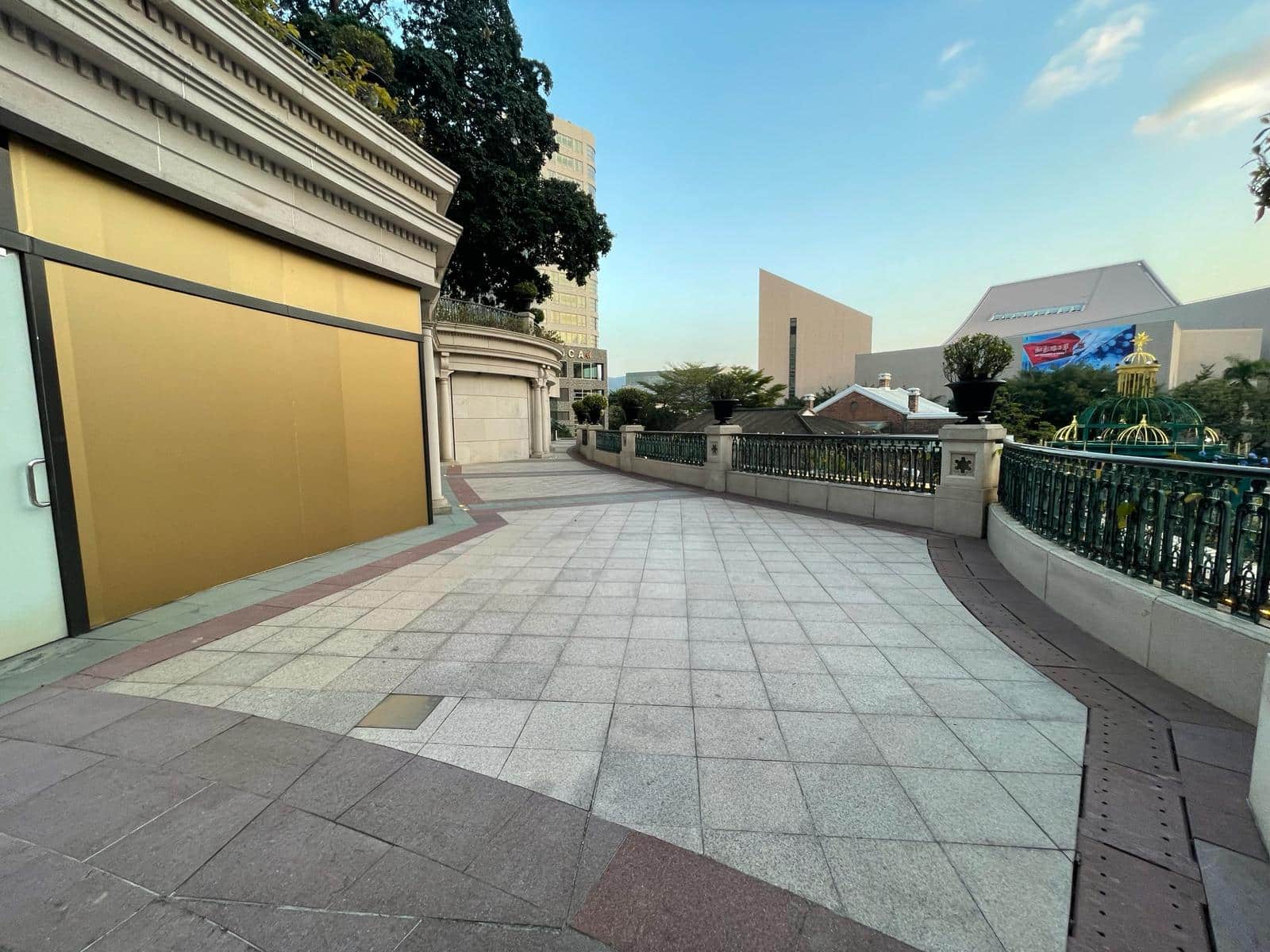 Ref#3013 features a wide tiled walkway curving beside a gold-walled buildingperfect for restaurant or shop rentalwith trees and modern buildings visible in the background under a partly cloudy sky.