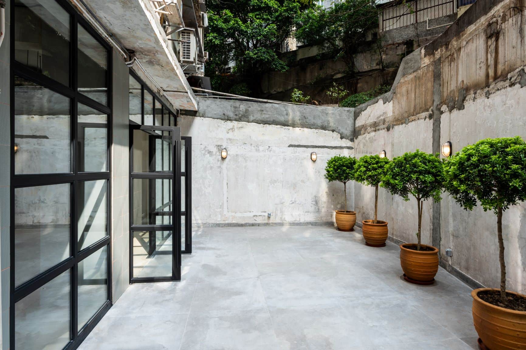 Concrete patio in Sai Ying Pun with potted trees beside a weathered wall, glass doors, and outdoor wall lights.