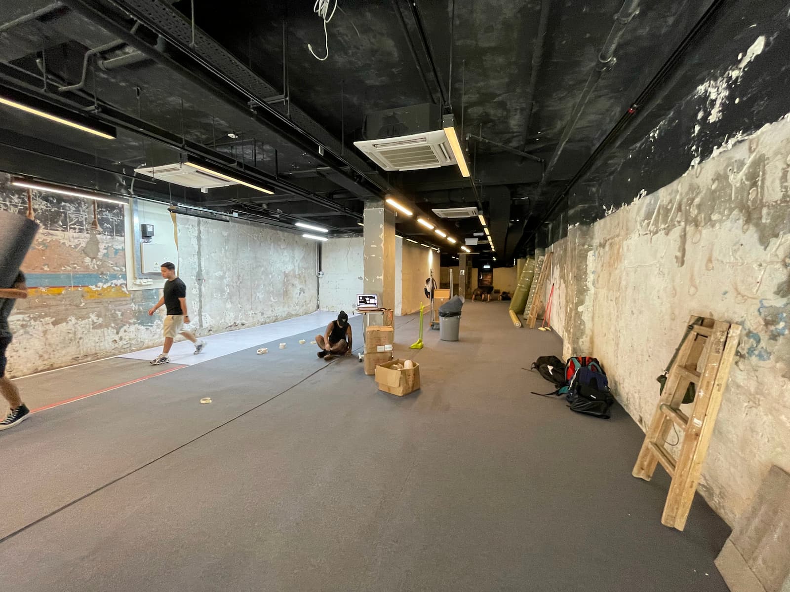Ref#3015 features a spacious, unfinished indoor area with exposed pipes and concrete wallsperfect for a shop or office. Several people are working, with boxes, bags, and a ladder scattered throughout the space.