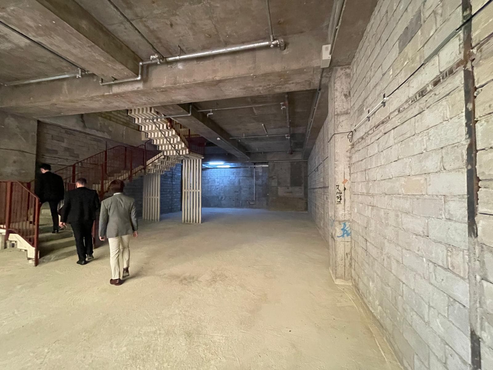 Four people in formal wear ascend and descend red stairs in Ref#3014, a spacious unfinished concrete basement with exposed pipes, dirt floor, and moody blue lightingideal for a unique restaurant rental or lease shop.