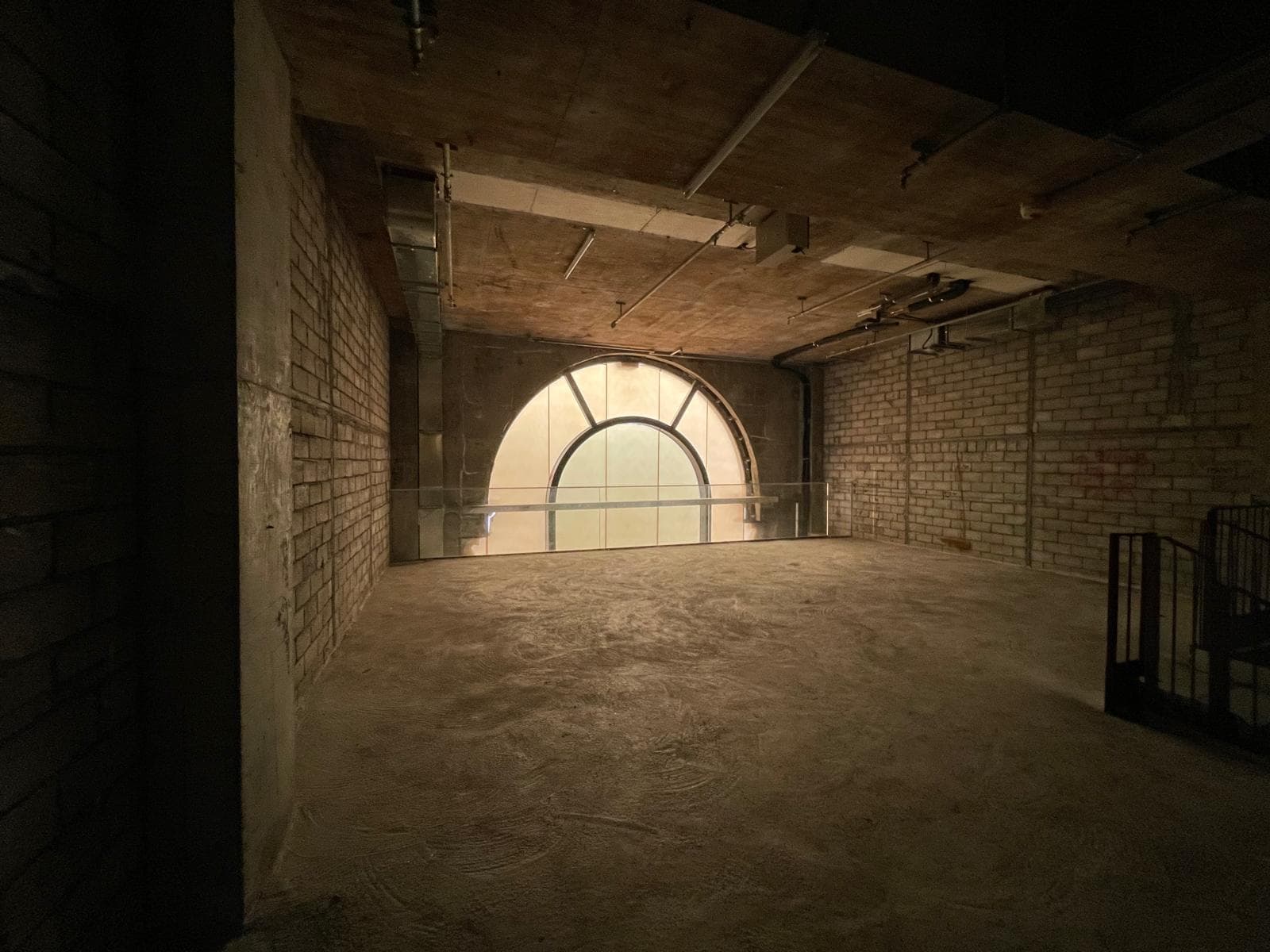 Ref#3014: This empty room features concrete floors and walls, a large arched window with hazy light, exposed piping on the unfinished ceiling, and a metal railingideal for use as an office or shop.