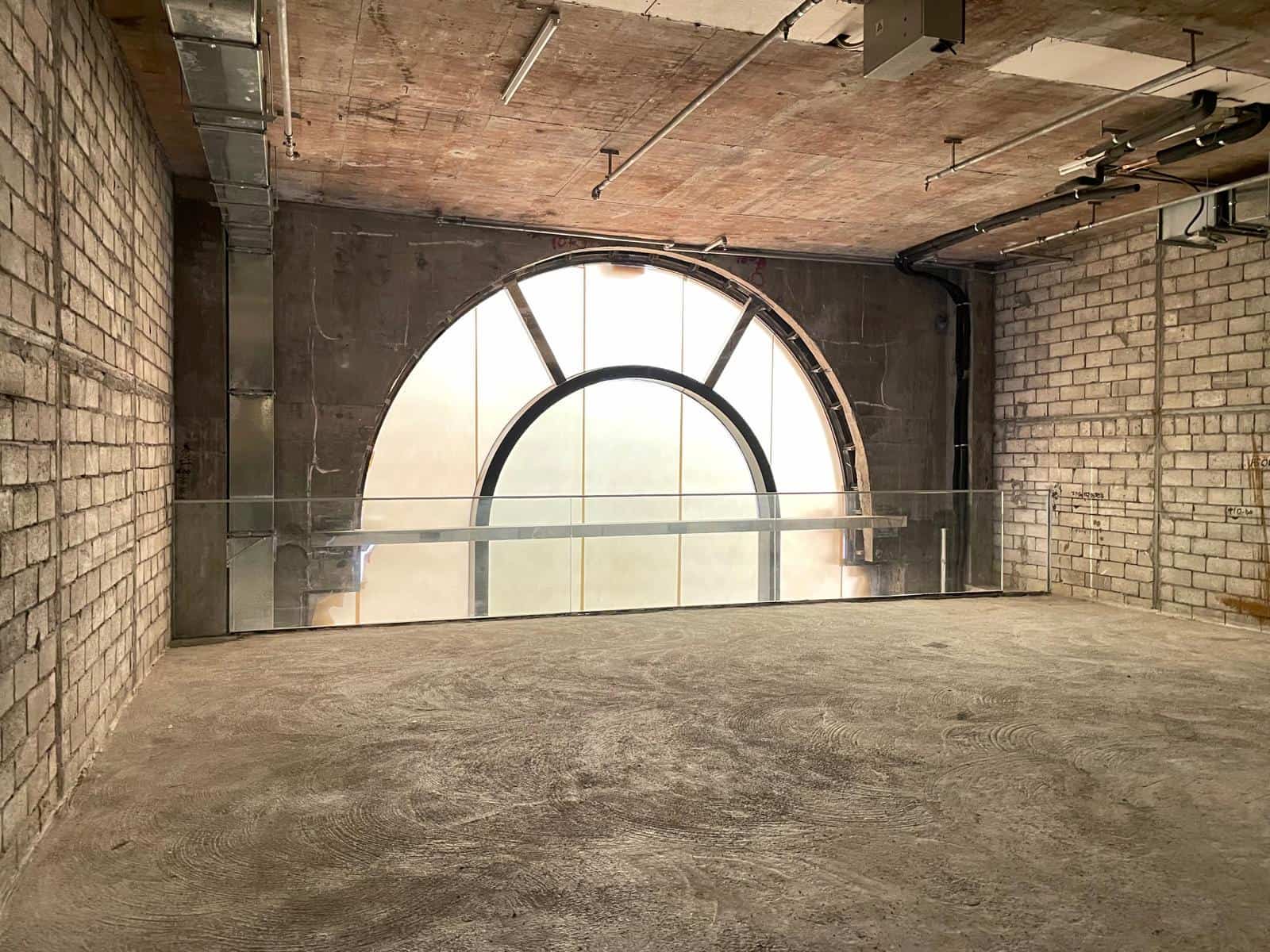 Ref#3014: Spacious industrial room with exposed brick walls, concrete floor, large arched frosted window, glass railing, and metal ceiling pipesideal to rent as an office or shop.