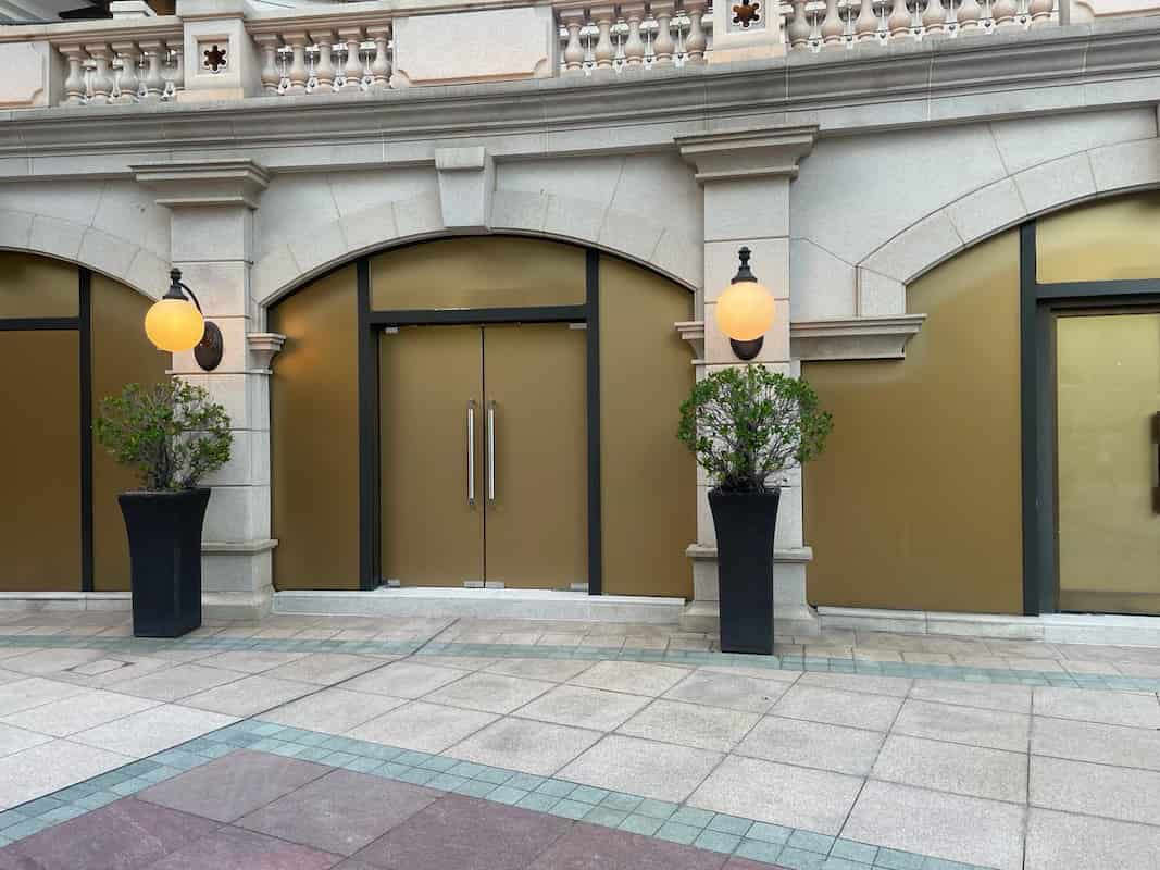 Ref#3012: A closed storefront with gold-covered windows, double glass doors, large potted plants, and globe wall lamps in a stone building with decorative railingideal for shop rental.