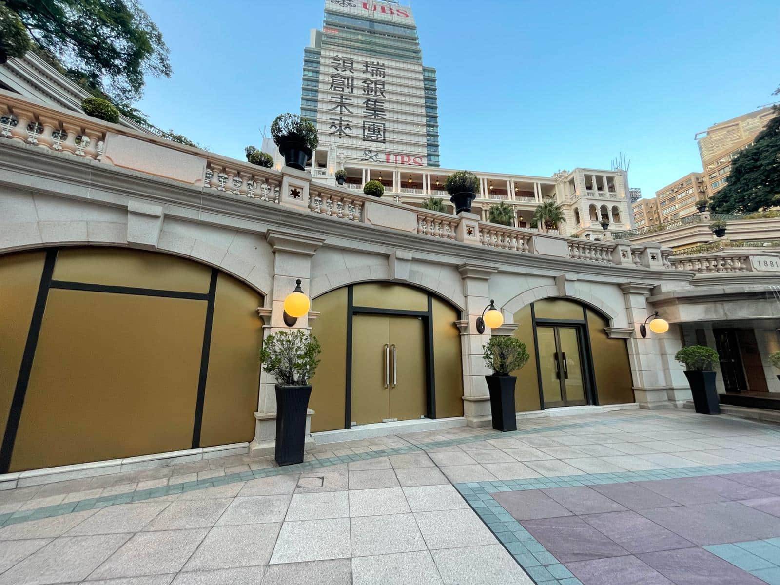 Ref#3010 features a modern facade with large gold-colored windows and doors, potted plants, and round lightsideal for office or shop rental. Behind it, a tall tower with English and Chinese signs rises against the blue sky.