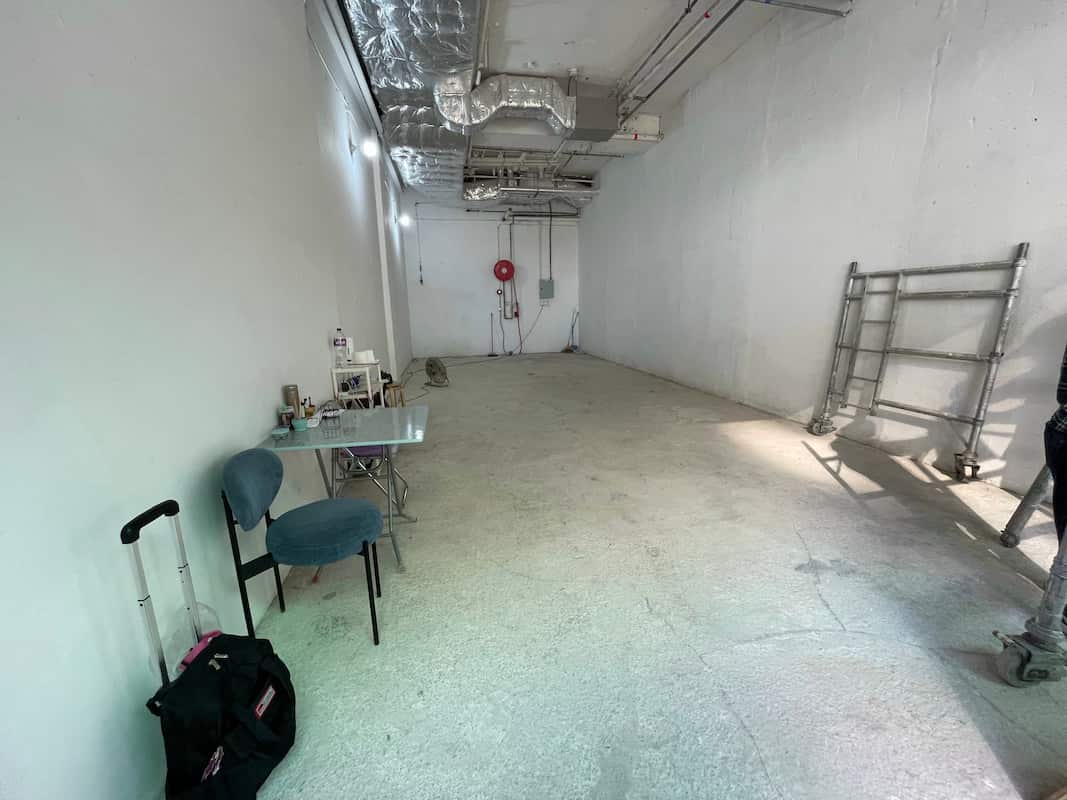 Ref#3009: An industrial-style shop rental with white walls, exposed ceiling pipes, a small table with two chairs, a suitcase, and black bag to the left, plus a metal scaffold on the right.
