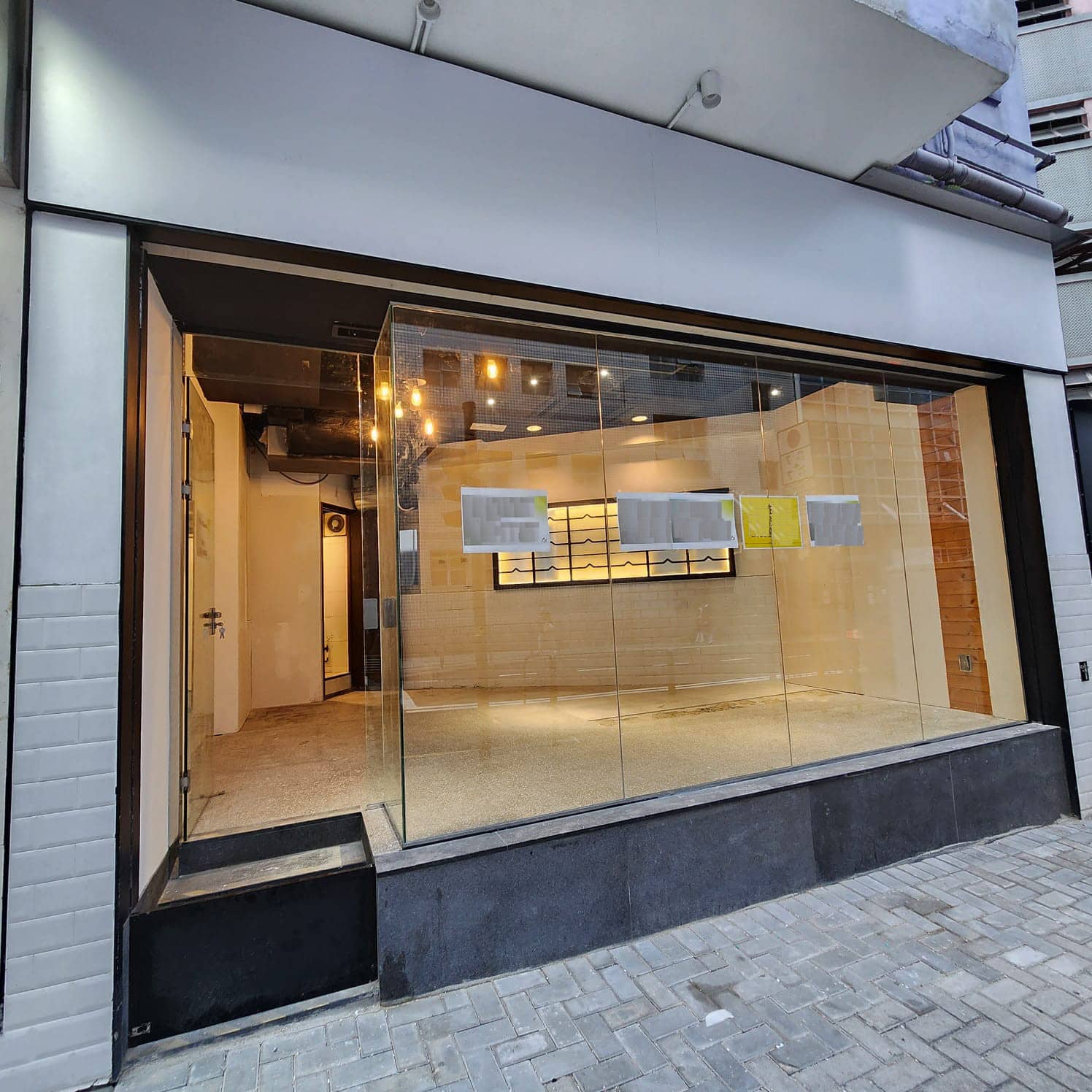 Ref#3008 is a modern, empty shop rental featuring large glass windows, light wood-paneled walls, ceiling lights, and papers on the window. The entrance has a small step up from the paved sidewalk.