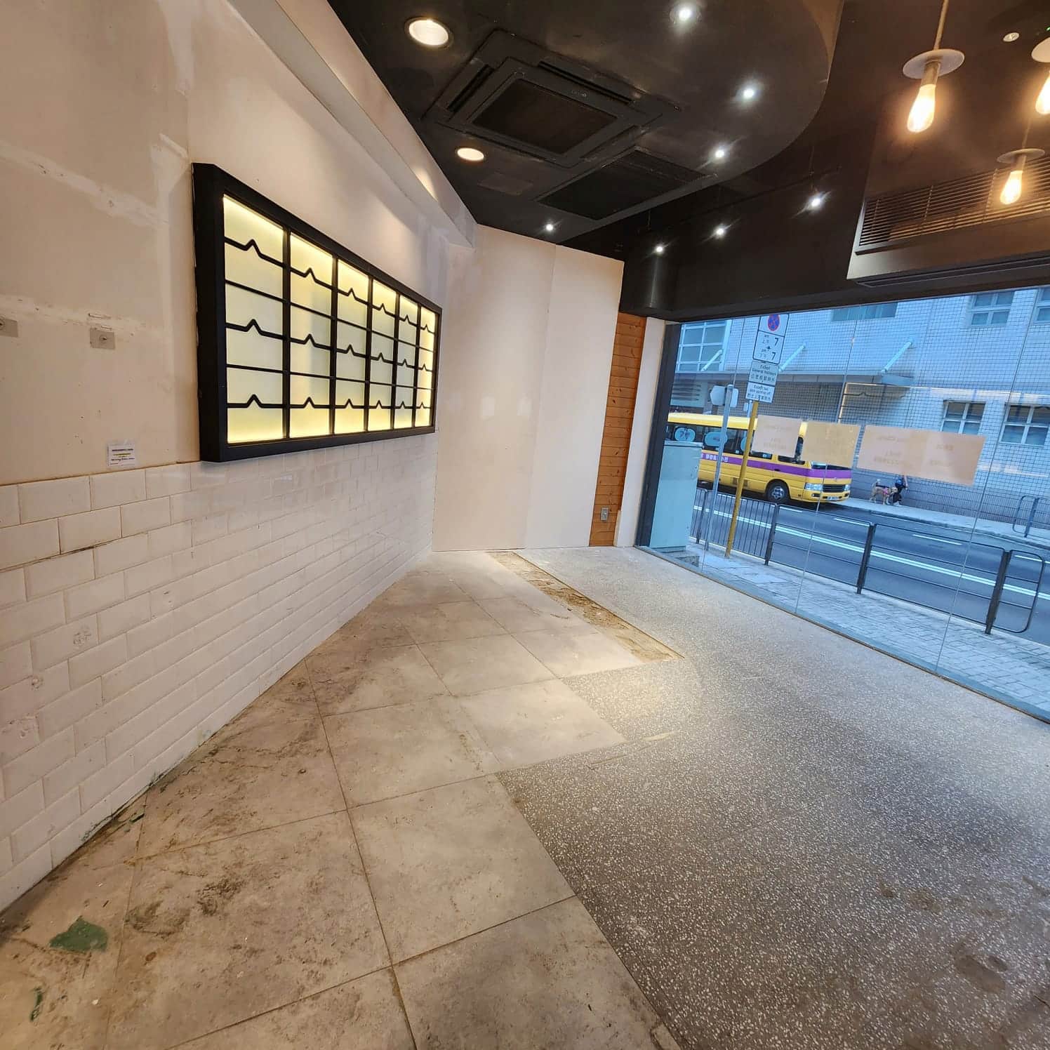 Ref#3008: Spacious store with white tiled walls, spotlights, large windows showing street and yellow bus, and a lit display case. Floor is partly tiled, partly unfinished 6ideal to rent as a shop or office.