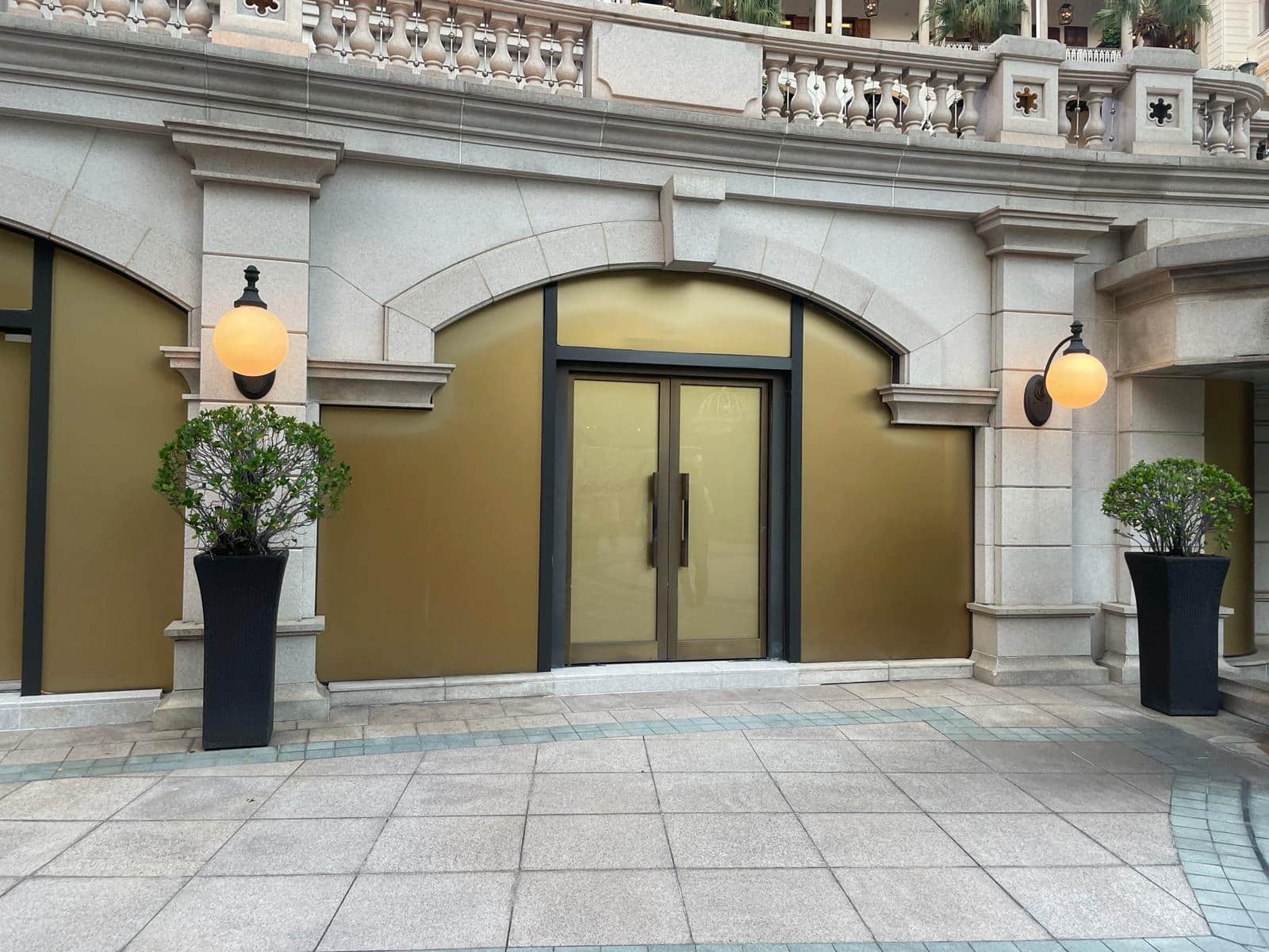 Ref#3010: A closed glass door with gold-tinted windows in a stone building, perfect for shop rental, flanked by two large potted plants and round wall lights on a tiled courtyard.