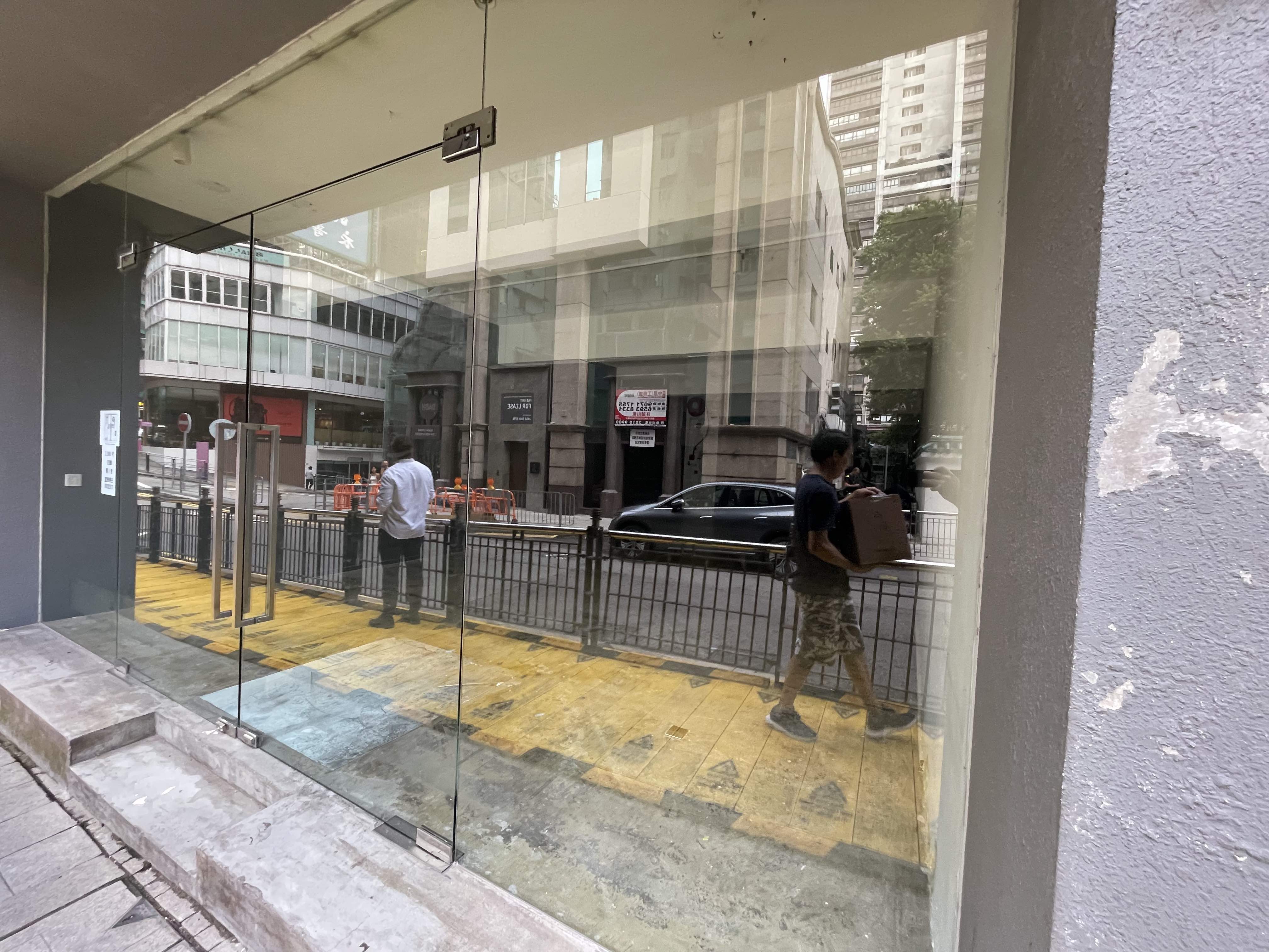 Two people walk past a glass-walled building, Ref#2986, one carrying a box. Across the street, city buildings and a black car are visible and reflected in the glass. They may be interested in renting office space.