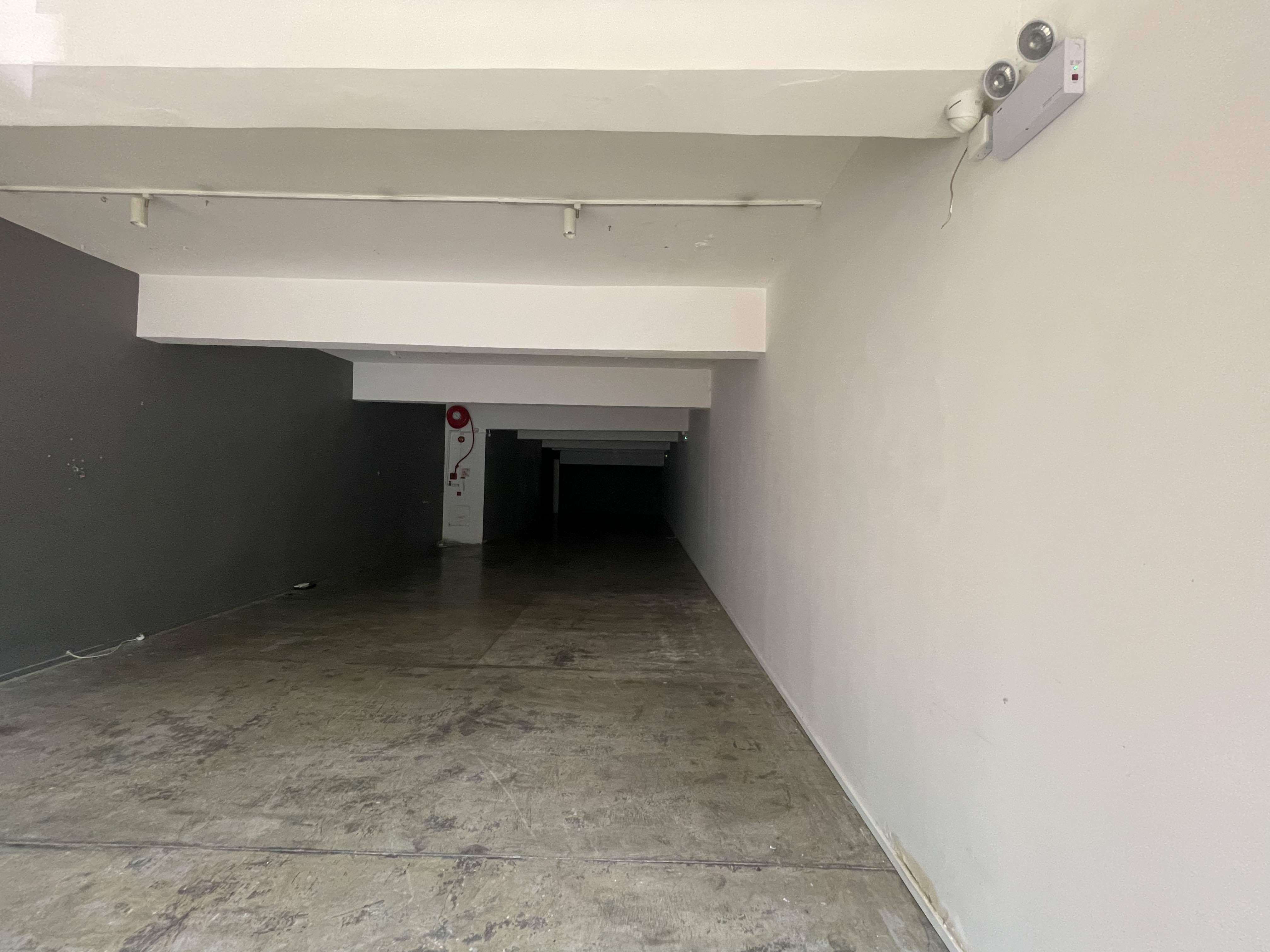 Ref#2986: An empty, dimly lit room with concrete floors, white walls, ceiling beams, and a fire extinguisher on a distant column 6ideal to rent as a shop or office. The space narrows and gets darker toward the back.