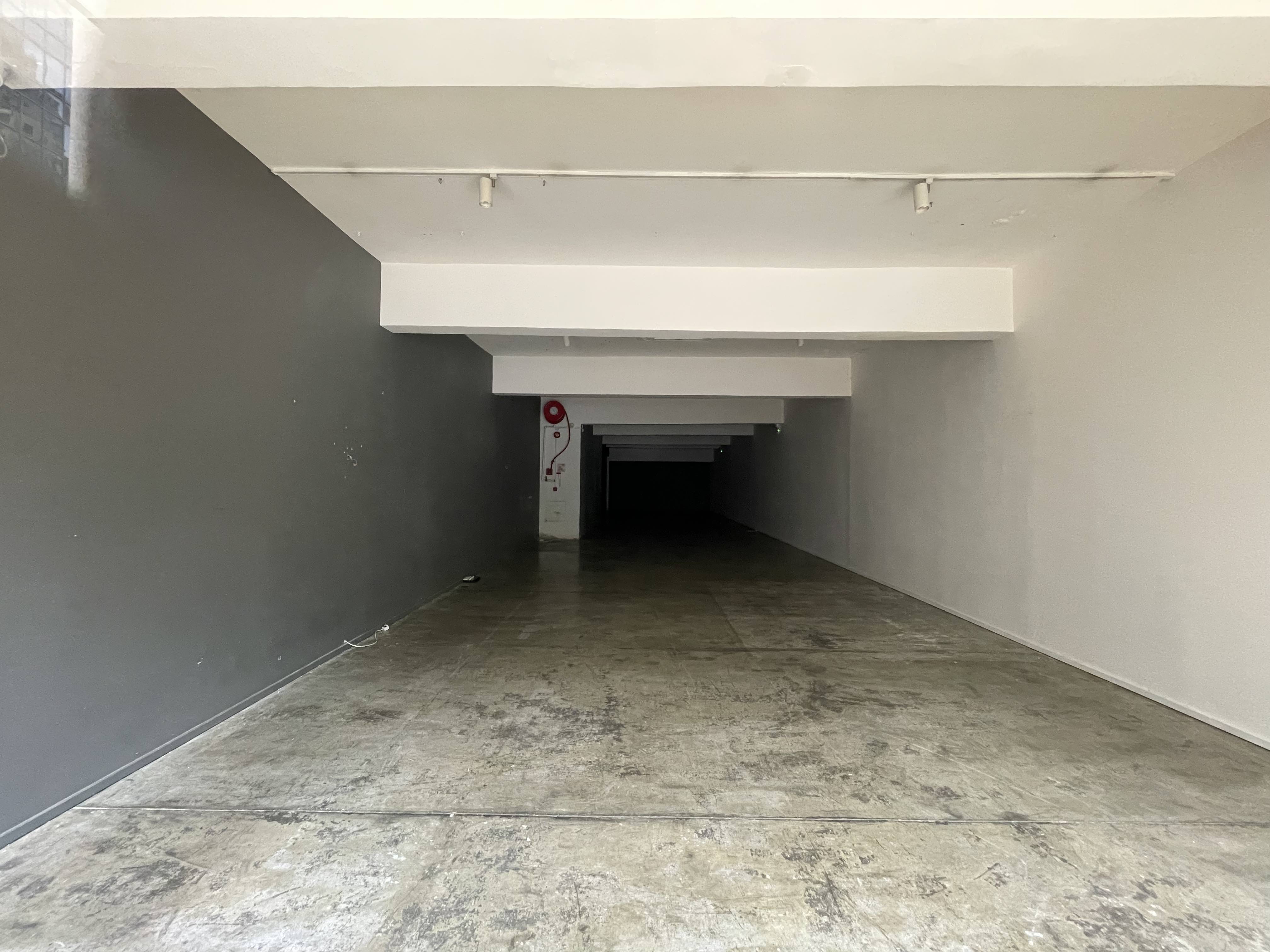 Ref#2986: An empty, narrow space with concrete floors, white ceiling beams, and gray and white walls 6ideal to rent for a restaurant, office, or shop. Undecorated; no furniture or windows; includes a fire extinguisher on the back wall.