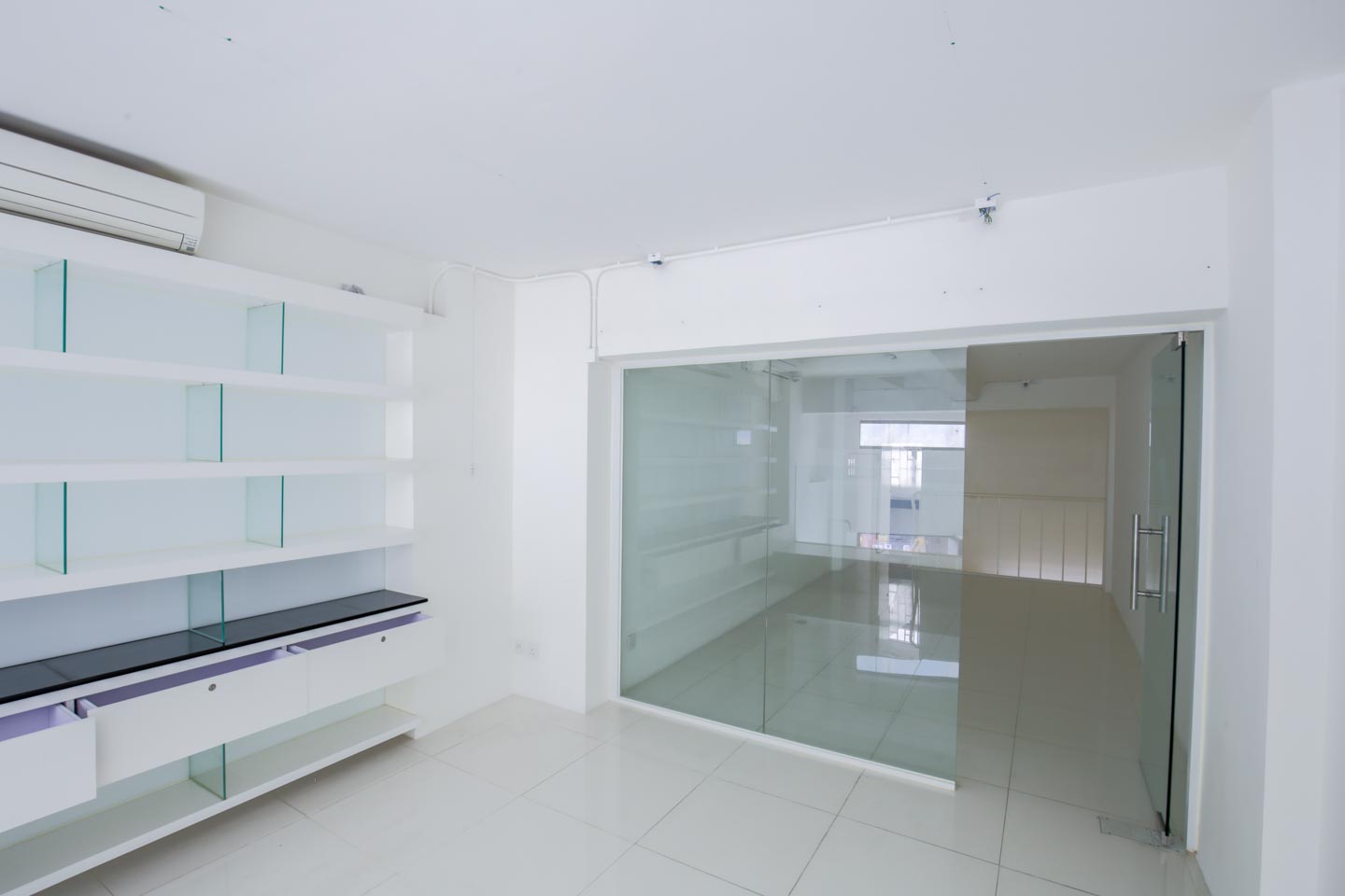 Ref#2981: Bright empty room with white tiled floors, white walls, glass shelving on the left, and a large glass door1 ideal to rent as a shop or office1 leading to another open space in the background.