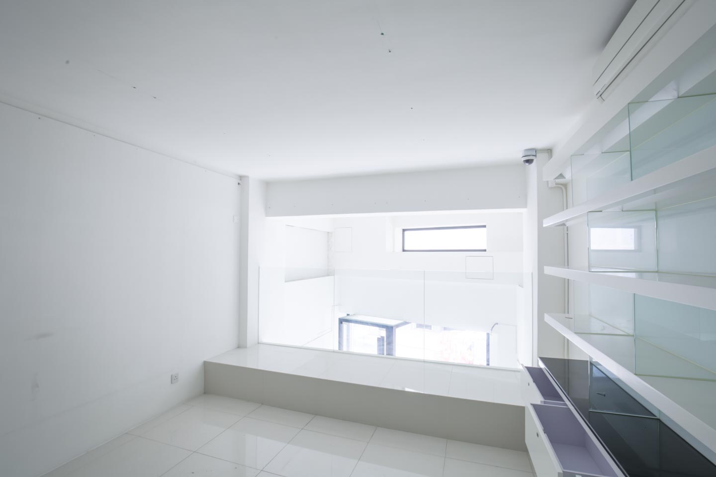 Ref#2981: A bright, modern empty room with white walls and floors, built-in glass shelves, glass railing, and a small rectangular window1 ideal for office rental with plenty of natural light.