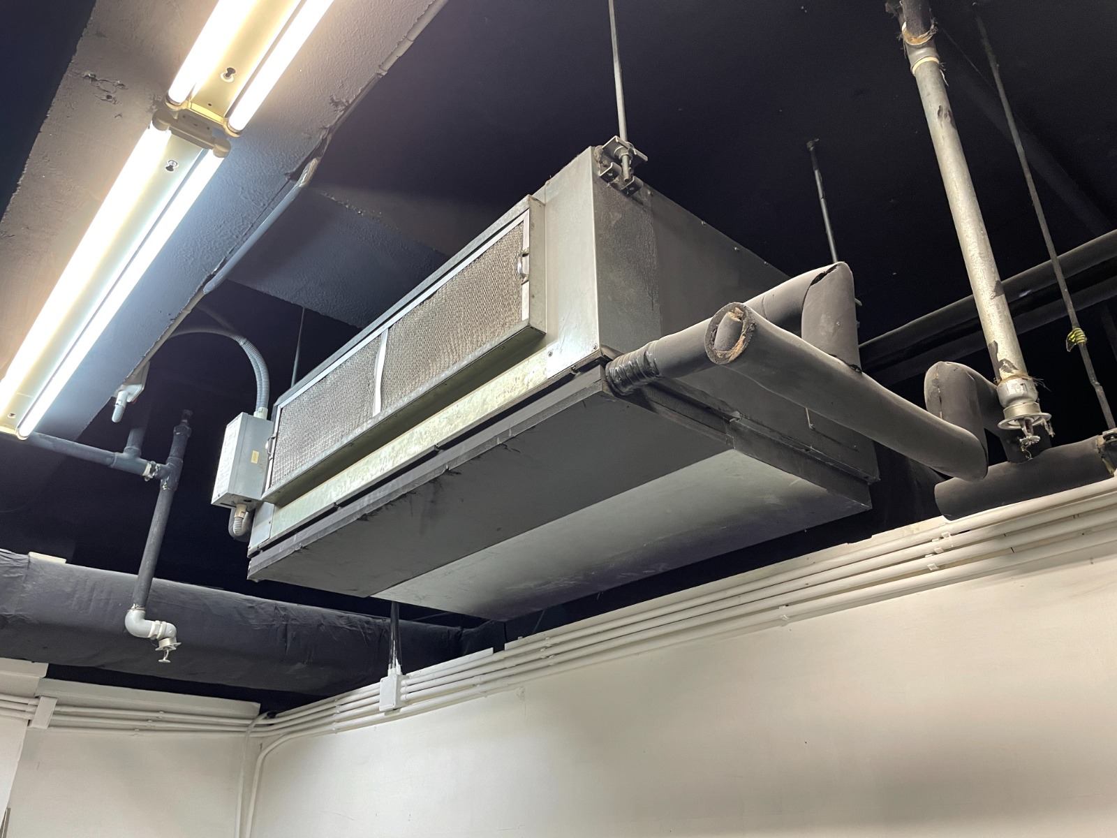 Ref#2982: A large metal HVAC unit is ceiling-mounted with exposed pipes and conduit, ideal for shop or office rentals. The black ceiling and pipes contrast the white walls, while fluorescent lights ensure bright illumination.