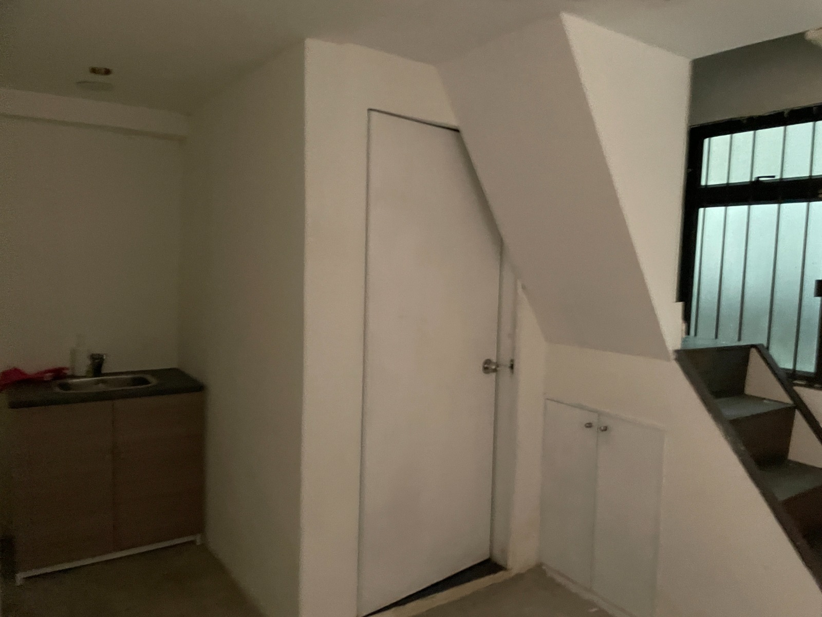 Ref#2982: Compact, dim room with a kitchenette left, white door center, stairs right, barred window at back. Suitable for office or shop rental. Storage cabinets built under stairs.