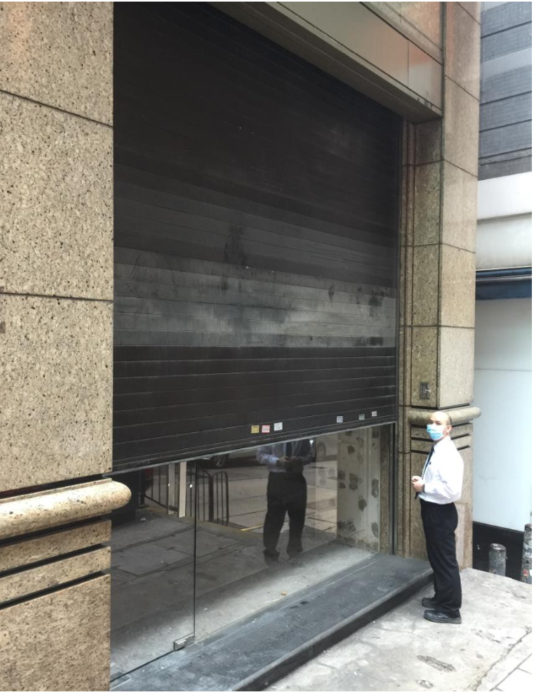 A man in a white shirt, black pants, and mask stands by a partially open black metal shutter on a city street beside Ref#2982, possibly checking out prime space to rent. His reflection is visible in the glass below the shutter.