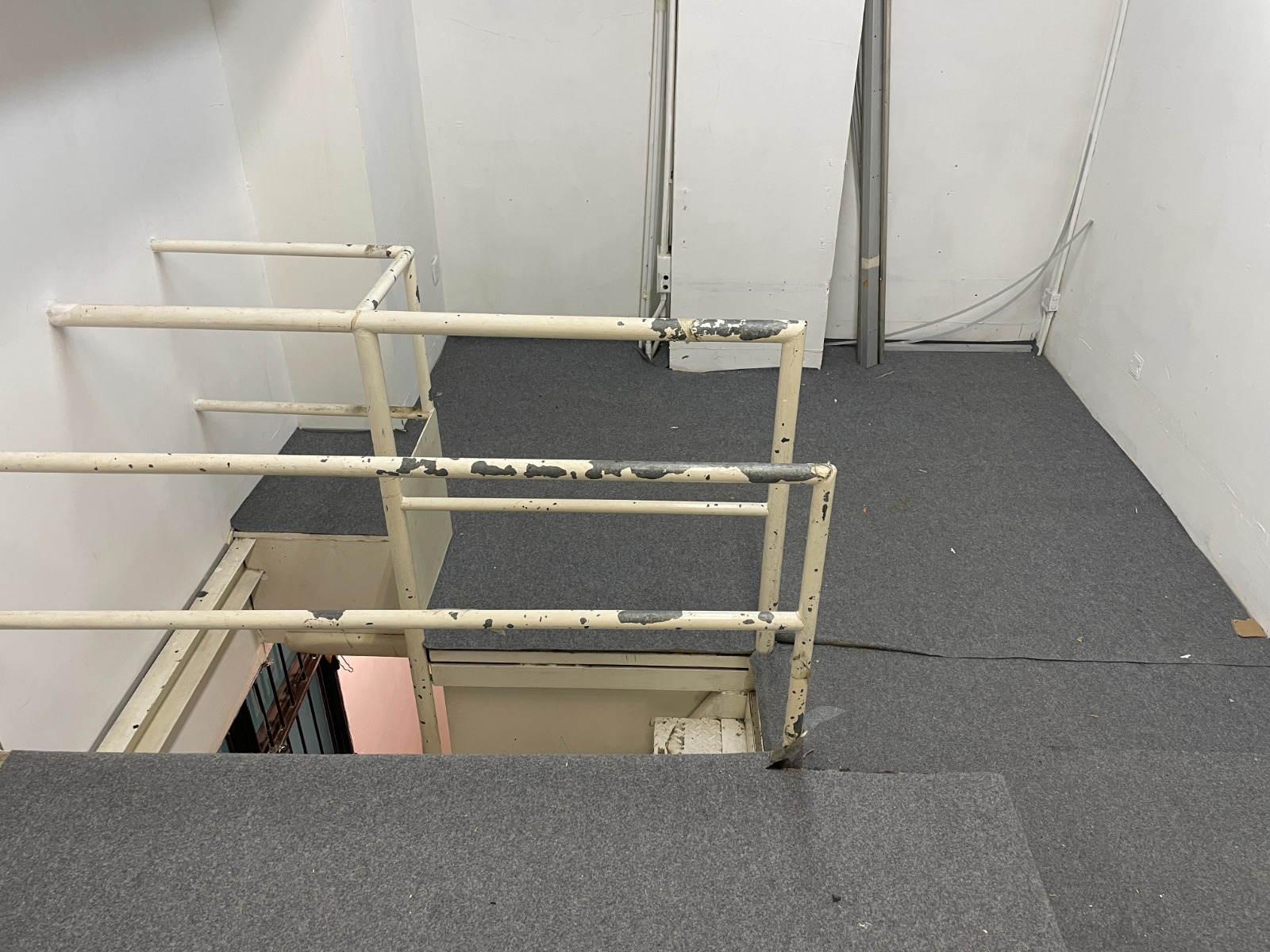 Ref#2982 features a carpeted staircase with chipped white metal railings leading to a lower level40an unfinished area perfect for renting as an office, with exposed pipes and loose cables adding character to the blank white walls.